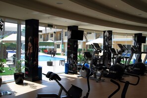 Et fitnesscenter med træningsudstyr, store vinduer og udsigt til en pool og bygninger.