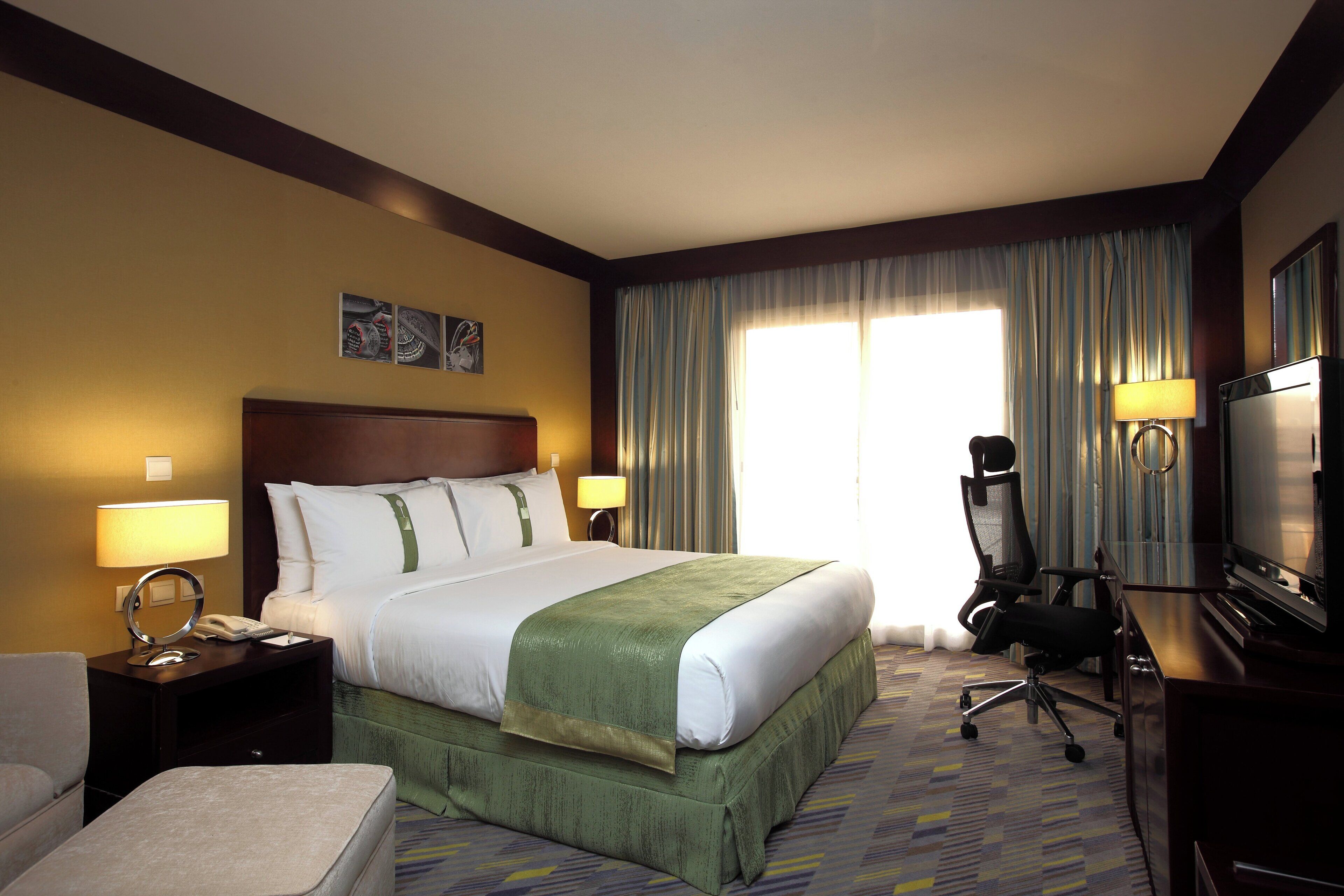 A hotel room with a large bed, a desk with a lamp, a chair, a television, and a window with curtains.