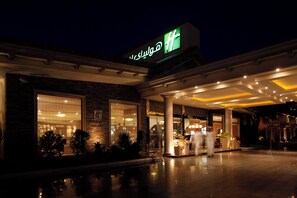 A hotel entrance with a large illuminated sign, a covered outdoor area, and a reflection on the ground. - Holiday Inn Al Khobar - Corniche by IHG (Al Khobar)