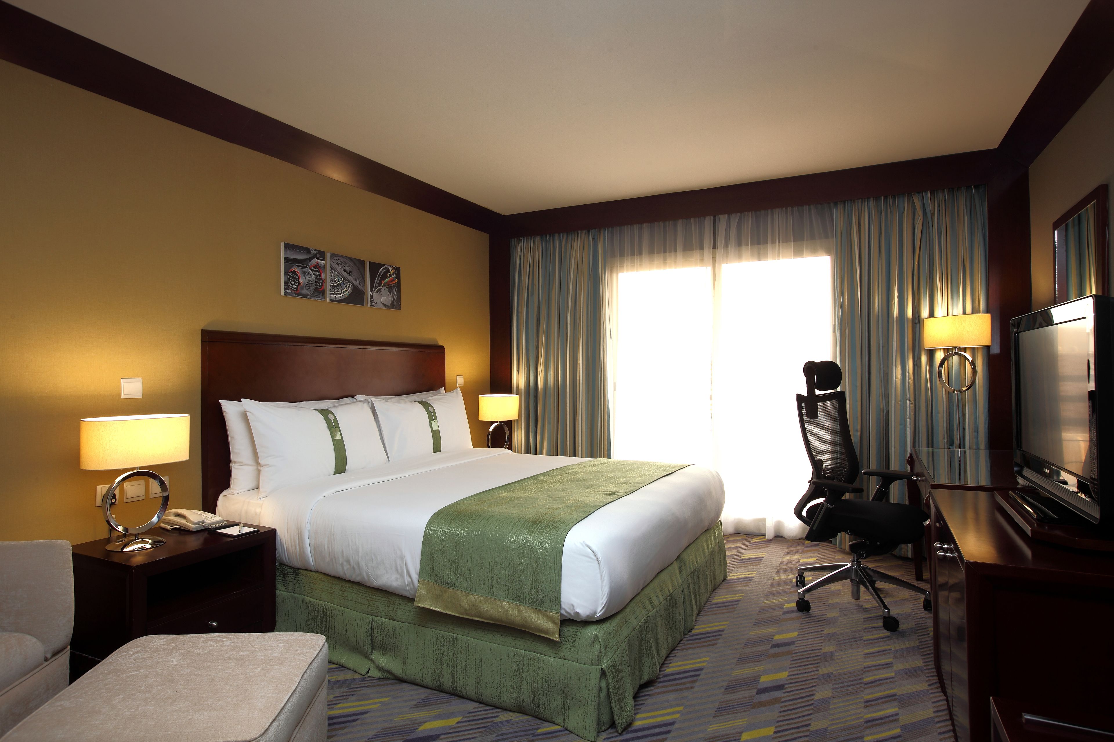 A hotel room with a large bed, a desk with a television, a chair, and a desk lamp.