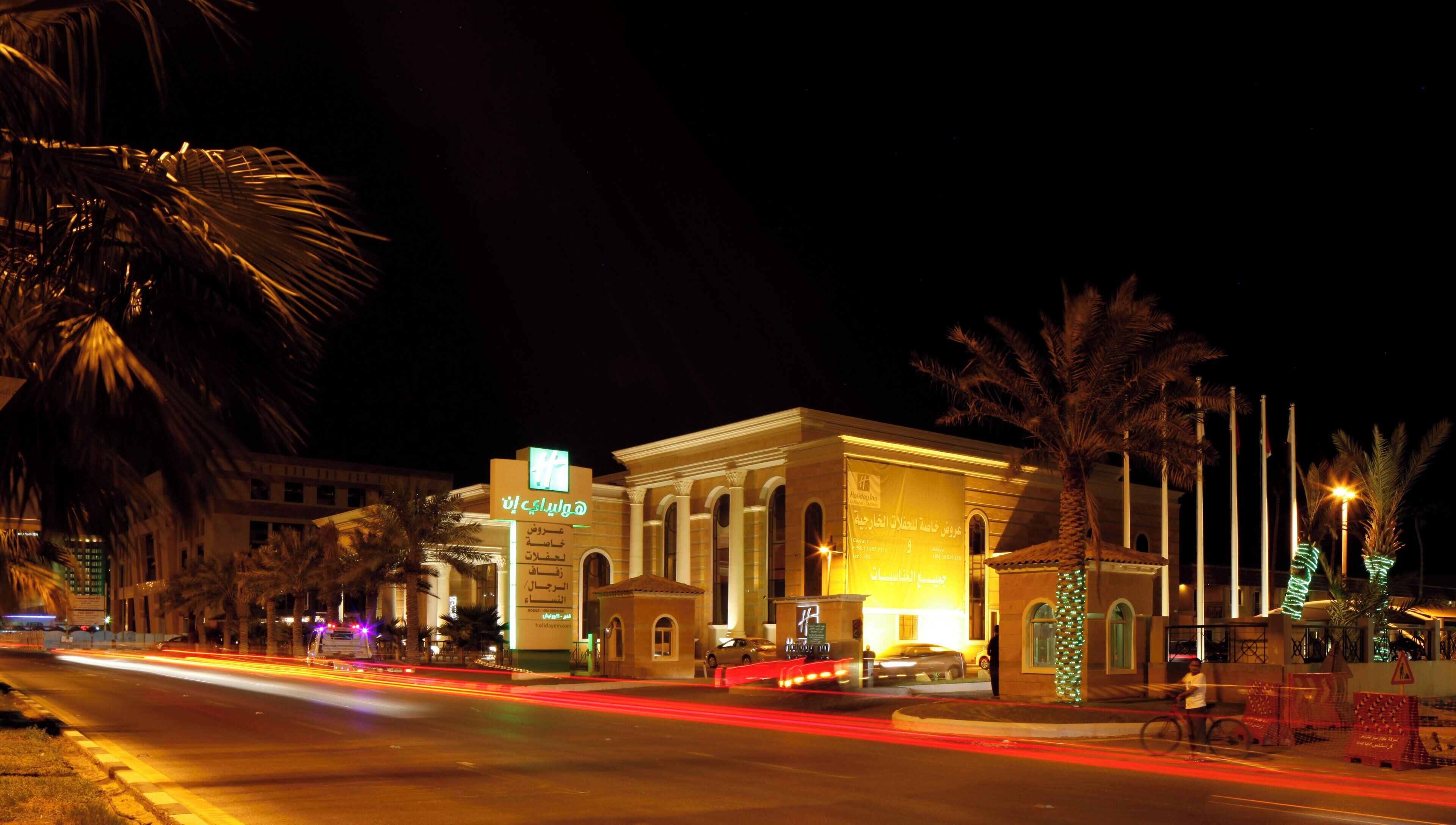 A hotel with a classical facade, palm trees, and illuminated signage.