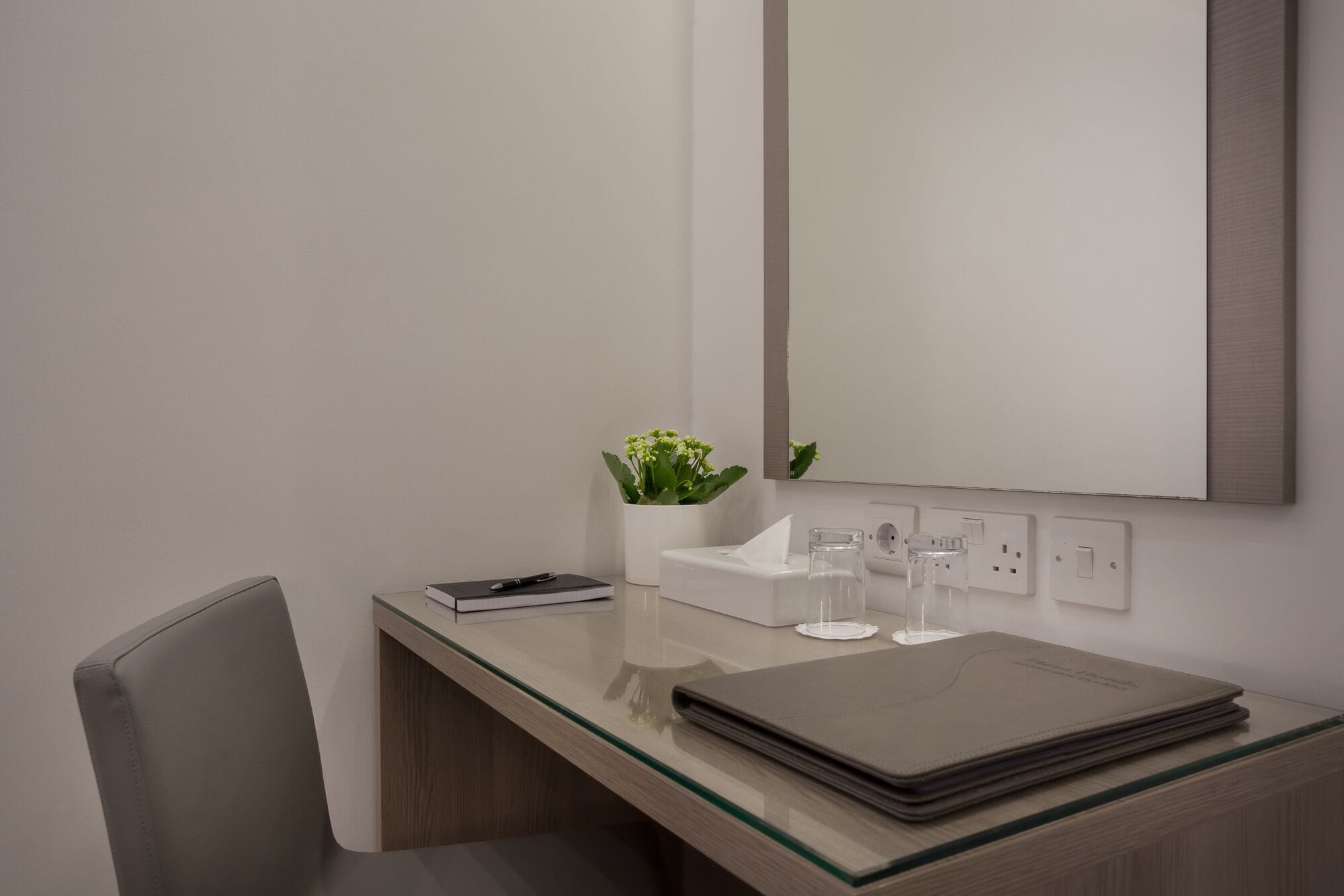 A modern bathroom with a glass countertop, a white sink, a mirror, a potted plant, and a stack of books.