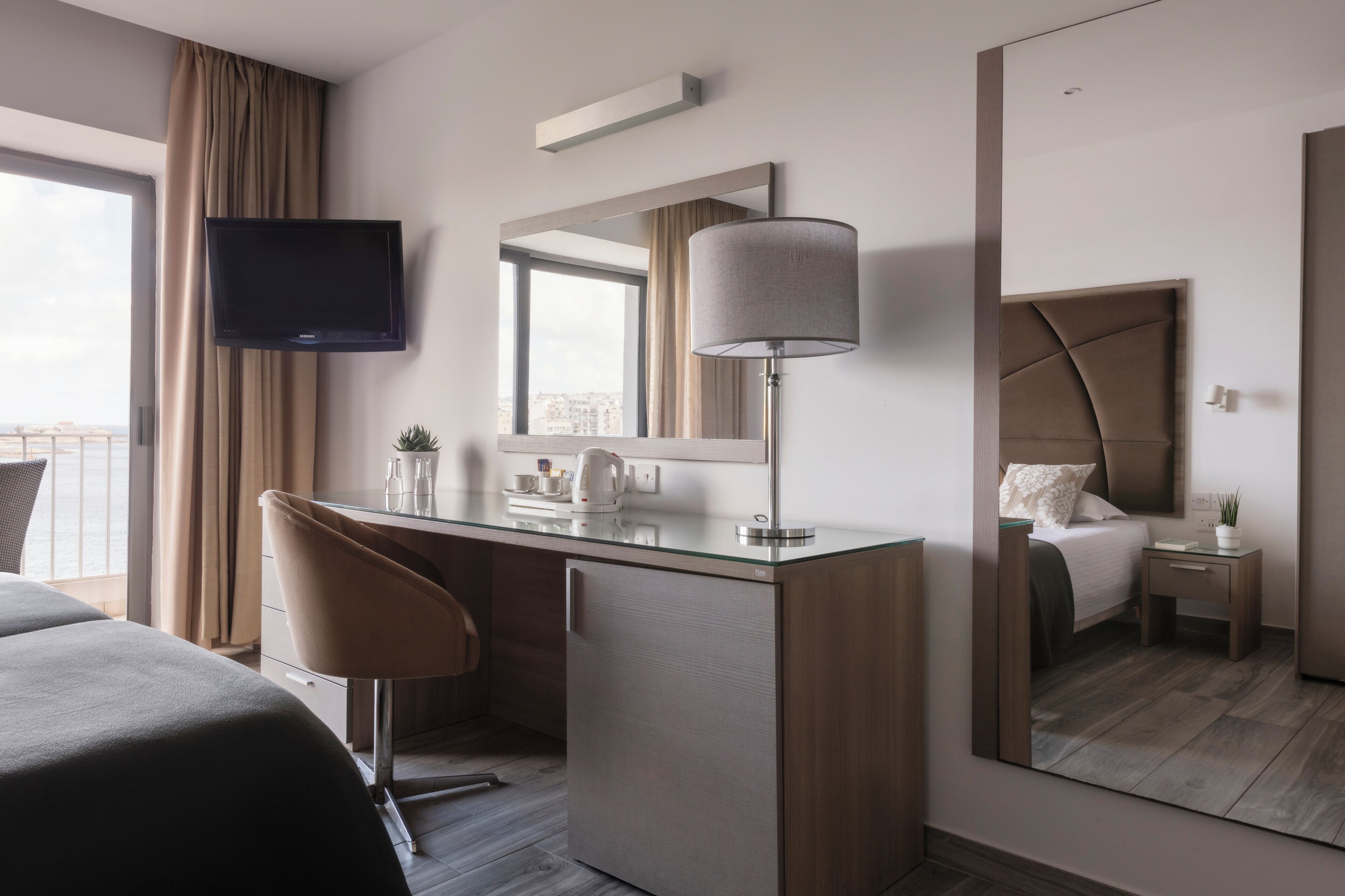 A hotel room with a bed, a desk with a chair, a TV, a lamp, a mirror, and a window with curtains.