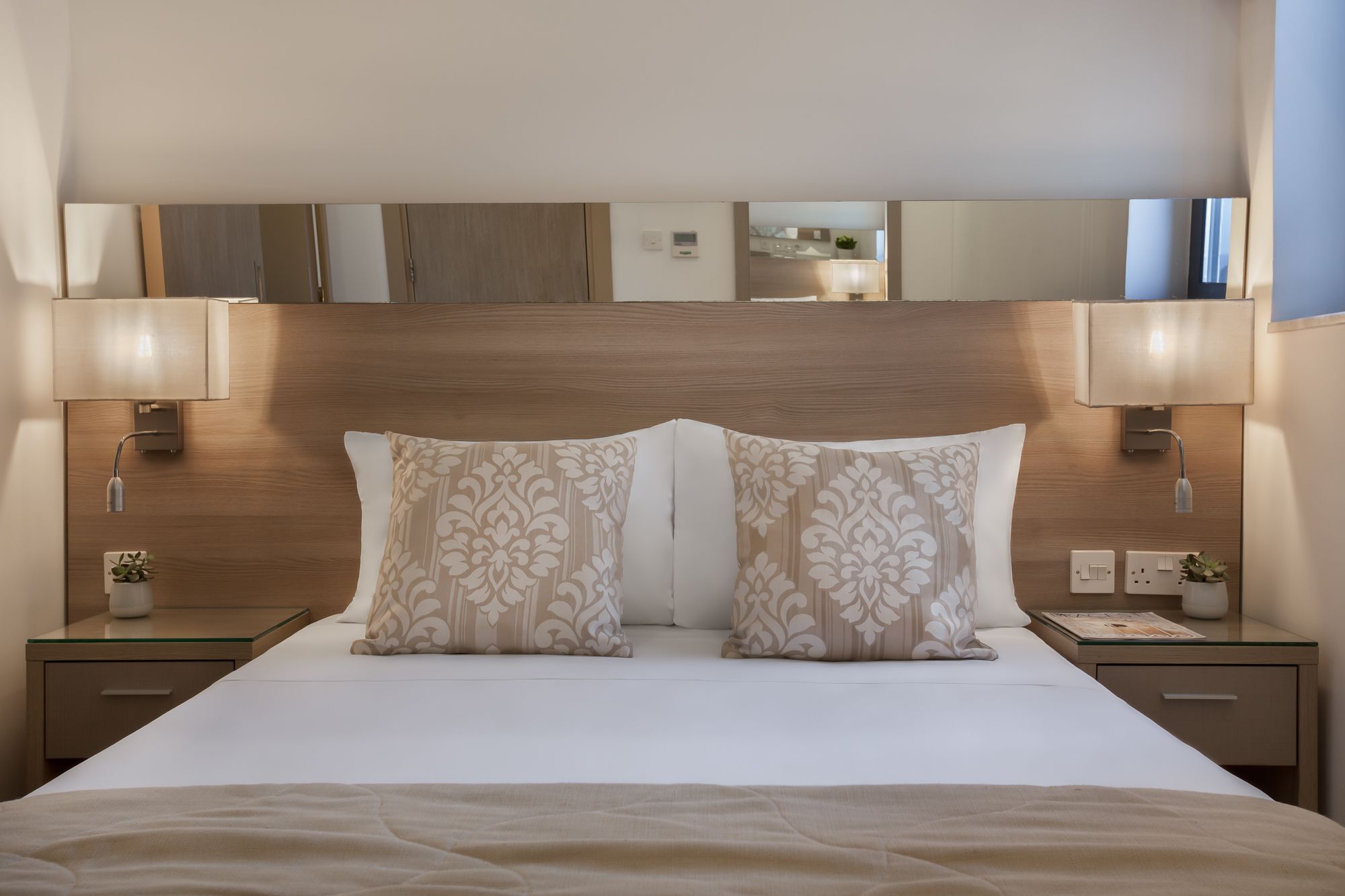 Habitación de hotel con una cama grande, dos mesitas de noche, un aparador, un espejo sobre la cama y una lámpara de pared.