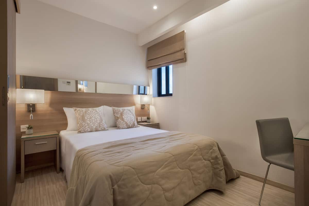 A modern hotel room with a large bed, bedside lamps, a small table with a plant, a chair, and a window with blinds.