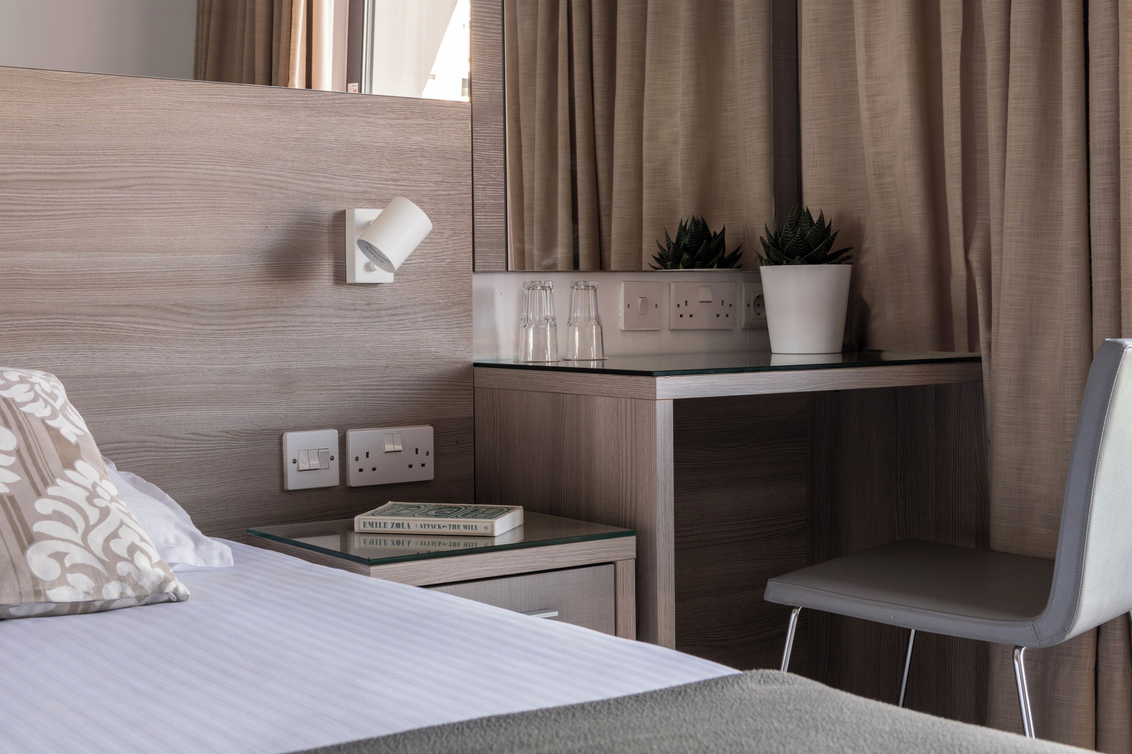 A modern hotel room with a wooden headboard, a glass desk with a potted plant, a white chair, and a bedside table with a book and glasses.