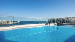 Piscina infinita en la azotea con agua azul cristalina, rodeada de barandales de vidrio, con vistas a un paisaje urbano costero y al mar.