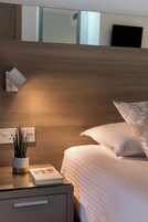 Una cama bien hecha con sábanas blancas y una sola almohada decorativa. Se ve una mesita de noche con un libro y una pequeña planta en maceta. La habitación tiene un cabecero de madera y una lámpara fijada a la pared.