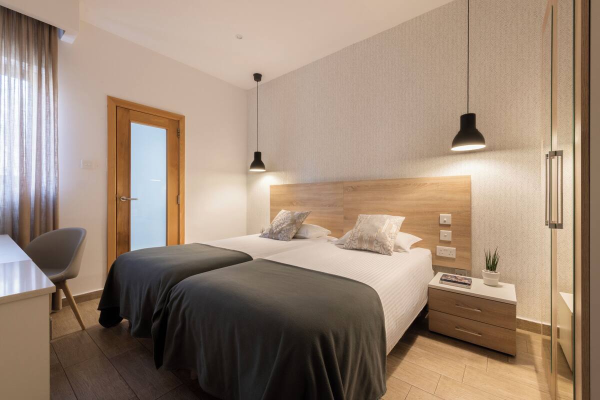 A modern hotel room with two beds, a desk, a chair, a bedside table with a lamp, and a small plant.