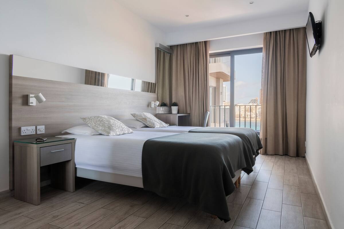 A modern hotel room with a large bed, bedside tables, a small desk, a potted plant, and a view of the city through large windows.