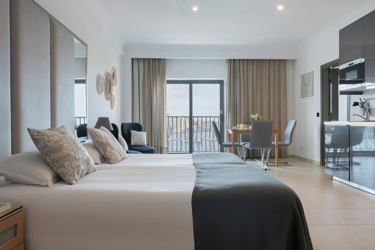 A modern hotel room with a large bed, a dining area, and a view of the city.