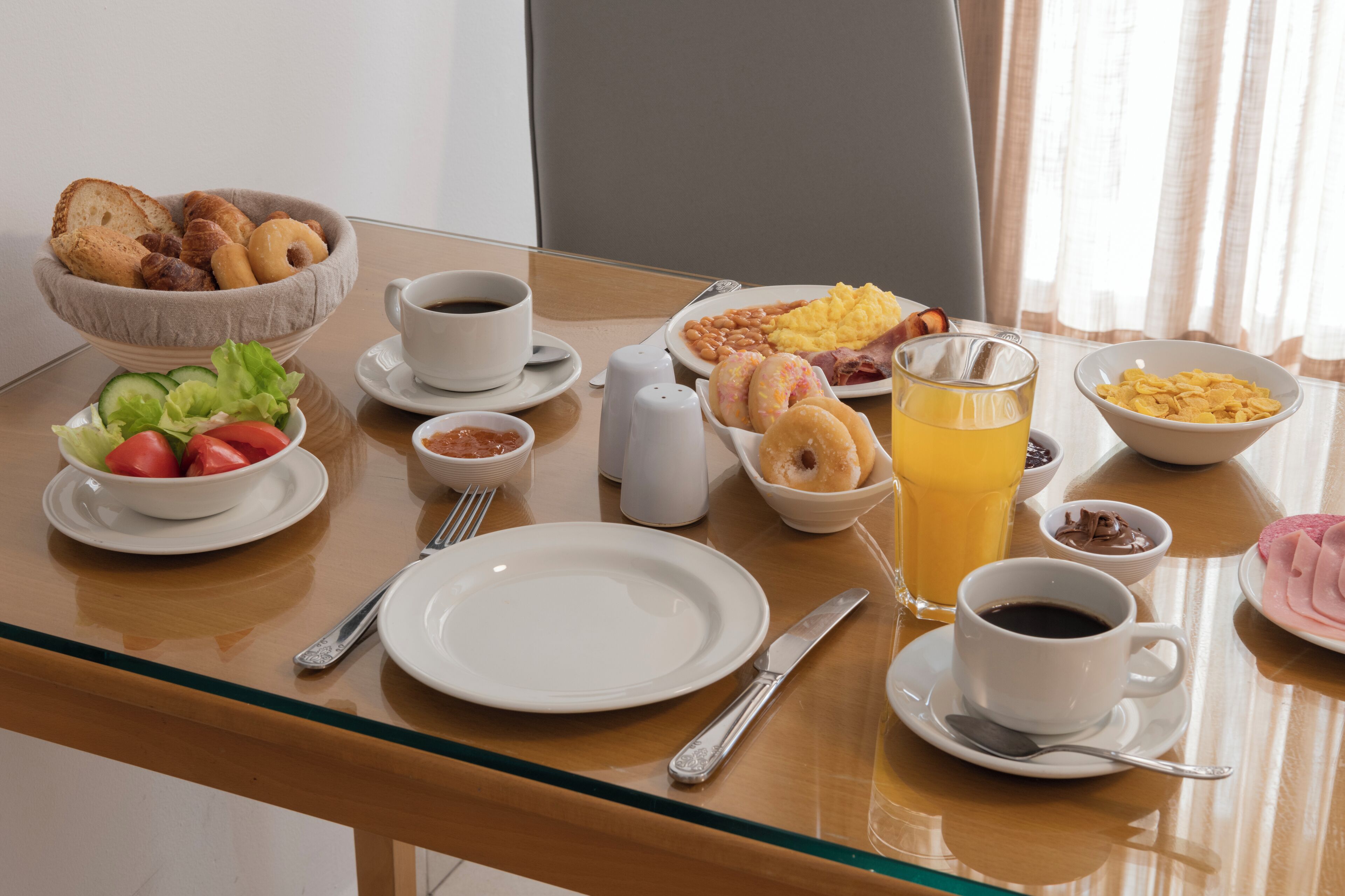 Una mesa de desayuno con diversos alimentos, tazas de café y un vaso de zumo de naranja.