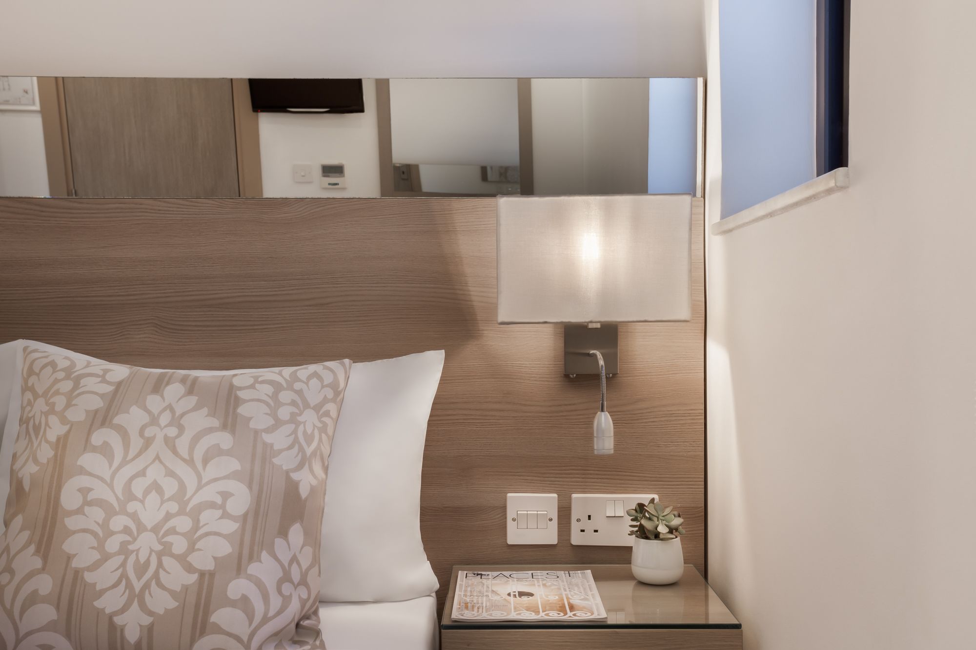 A hotel room with a wooden headboard, a bedside lamp, a small table with a plant, and a magazine.