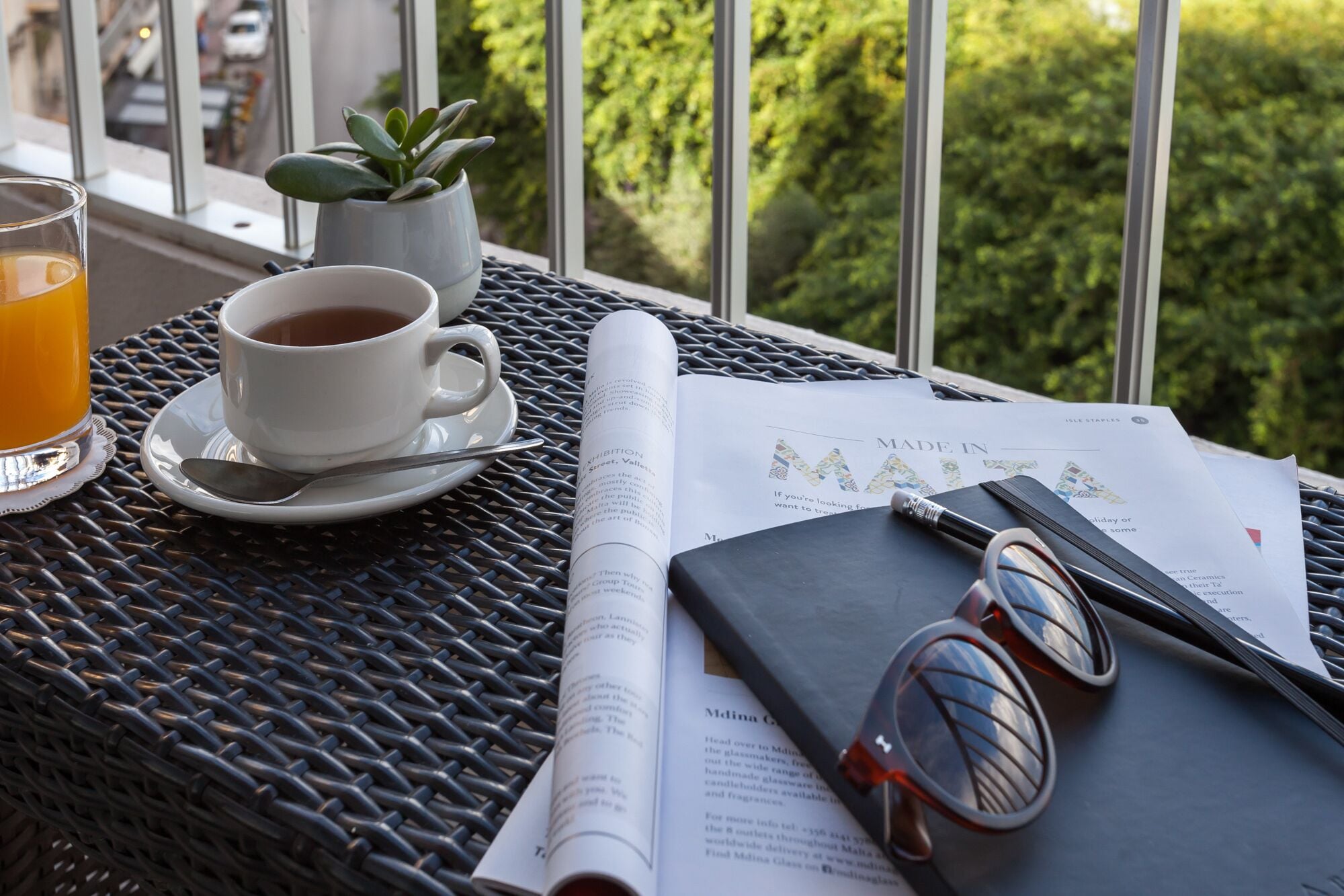 Una mesa con una taza de té, un vaso de jugo de naranja, un libro, un par de gafas de sol y una pequeña planta en maceta.