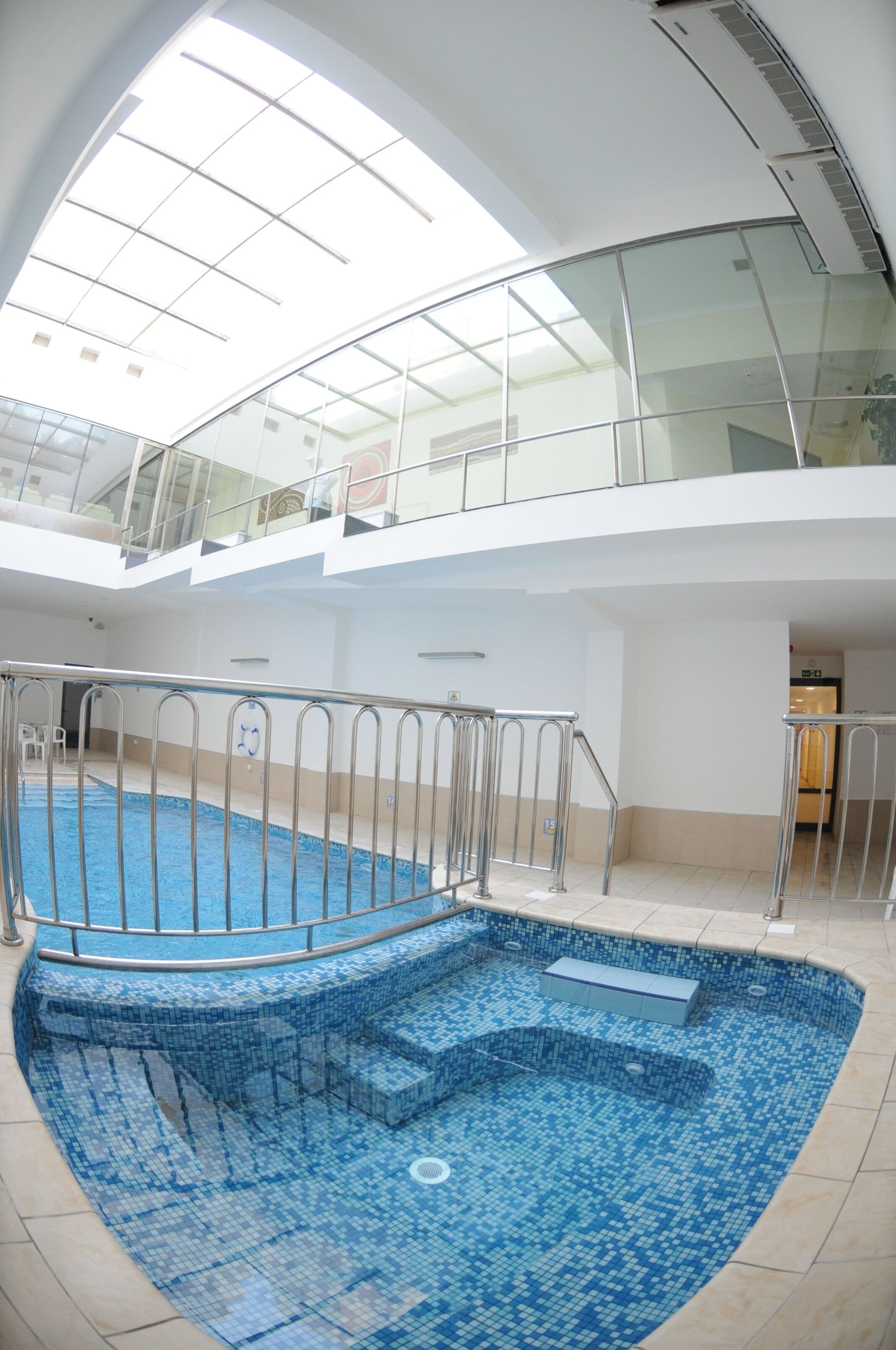 A large indoor swimming pool with a glass ceiling, surrounded by a metal railing, and a staircase leading down to the pool.