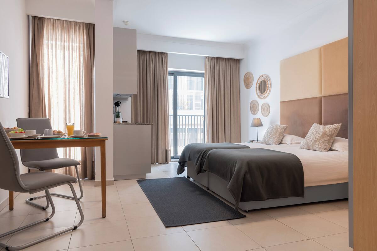 A modern hotel room with a bed, a desk, and a chair. There is a balcony with curtains and a view of the outside.