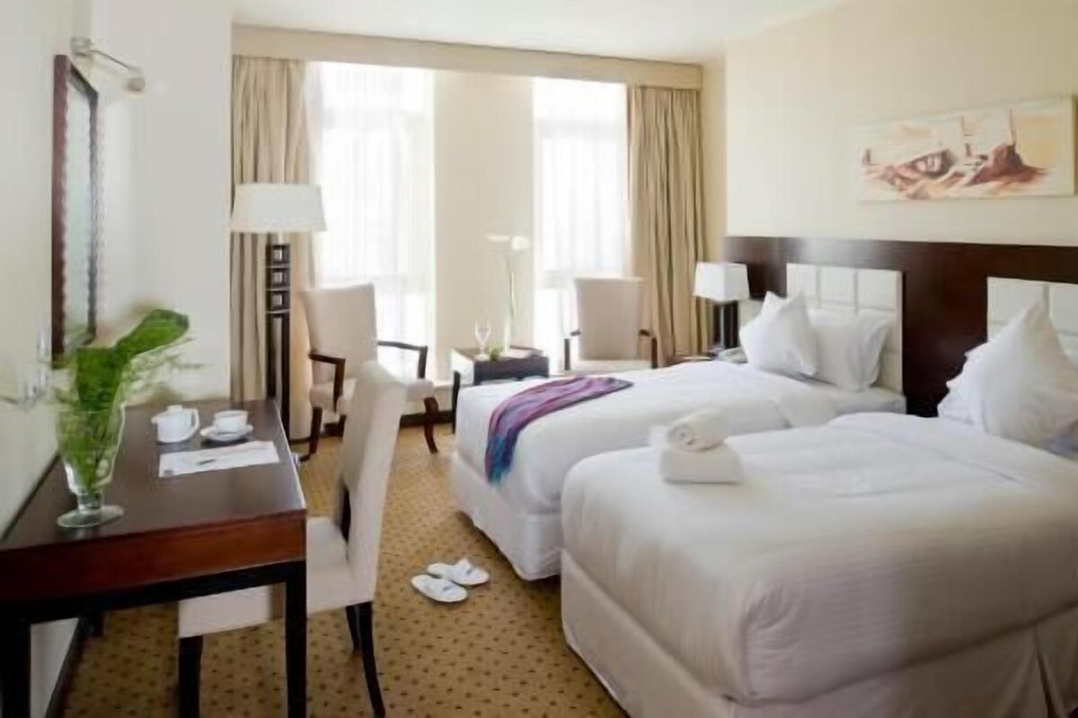 A hotel room with two beds, a desk with a chair, a lamp, and a painting on the wall.