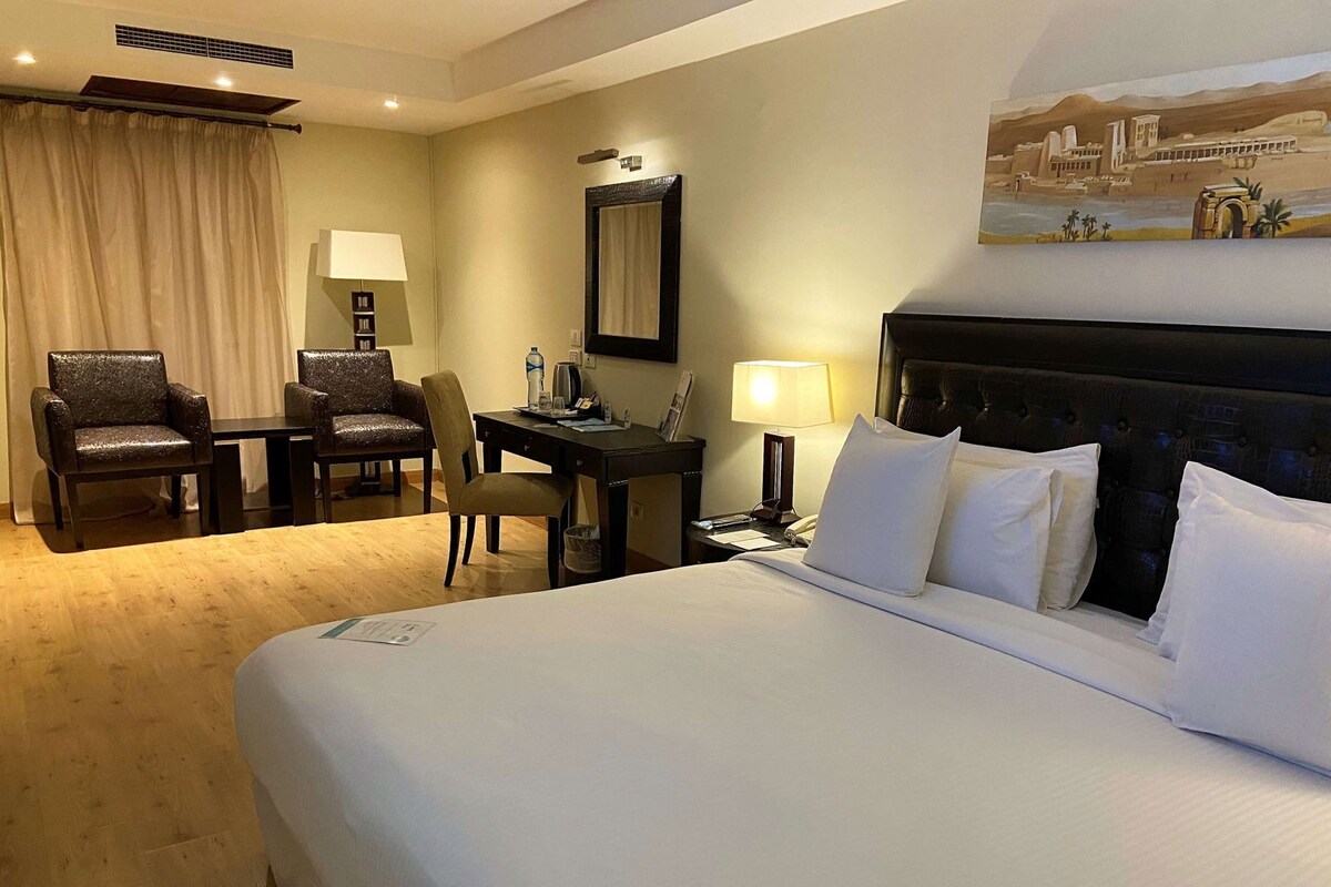 A hotel room with a large bed, a desk with a chair, a small table, and a lamp.
