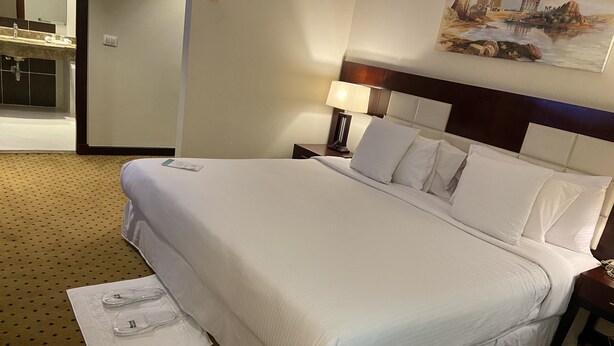 A hotel room with a large bed, a nightstand with a lamp, a painting on the wall, and a bathroom visible through an open door.