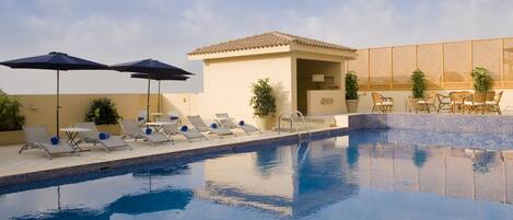 A swimming pool with lounge chairs and umbrellas, a small building with a tiled roof, and a wooden fence.