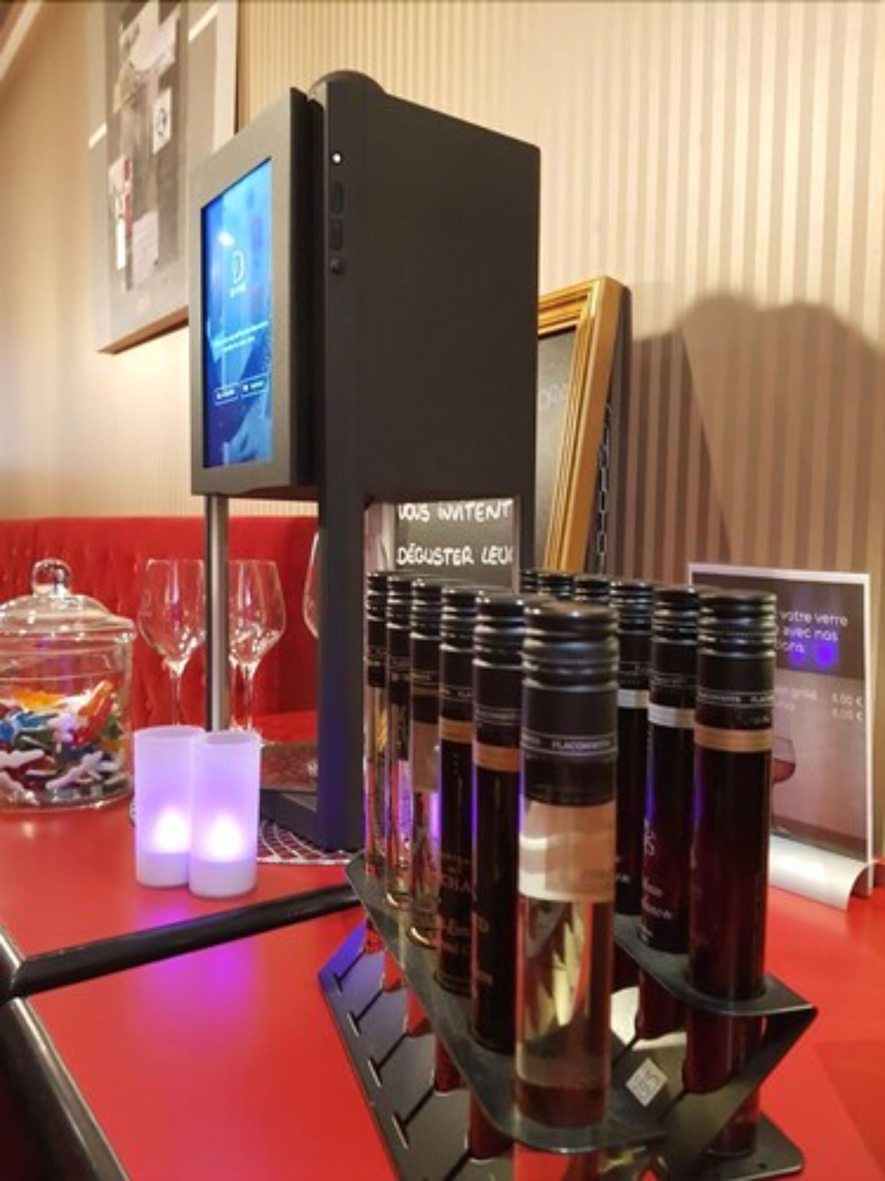 Uma área de bar com uma tela digital, seleção de garrafas e um recipiente de vidro com doces.