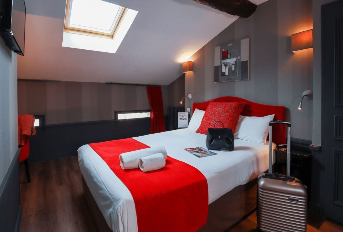 Ein Hotelzimmer mit Bett, Koffer, weißroter Bettdecke, Oberlicht und einer an der Wand befestigten Ablagefläche mit Gegenständen.