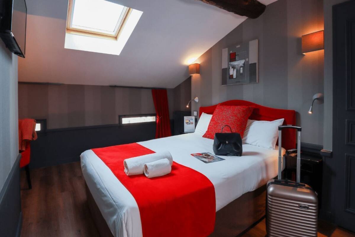 Una camera d'albergo con un letto, una valigia, una coperta a righe rosse e bianche, un lucernario e uno scaffale a parete con oggetti.
