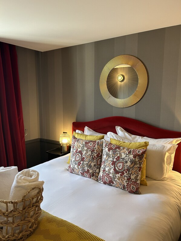 A hotel room with a neatly made bed, a decorative wall mirror, and a red curtain.