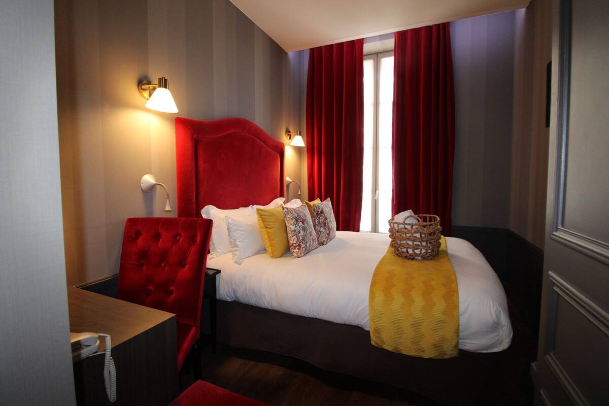 Una camera d'albergo con una testiera rossa, un letto con cuscini bianchi e decorati, un comodino con una lampada e un cesto di vimini sul letto.