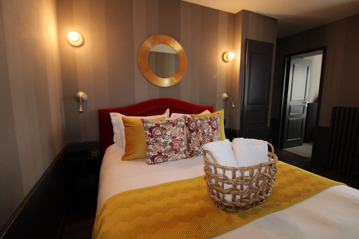 Camera d'albergo con un letto grande, una testiera rossa, cuscini decorati, una coperta gialla, un cesto di vimini con asciugamani e uno specchio rotondo appeso al muro.