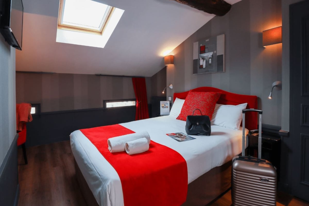 Una camera d'albergo con un letto, una valigia, una coperta a righe rosse e bianche, un lucernario e uno scaffale a parete con oggetti.