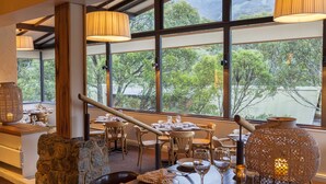 A restaurant with tables set for dining, large windows with a view of trees, and decorative lighting fixtures. - The Denman Hotel (Thredbo)