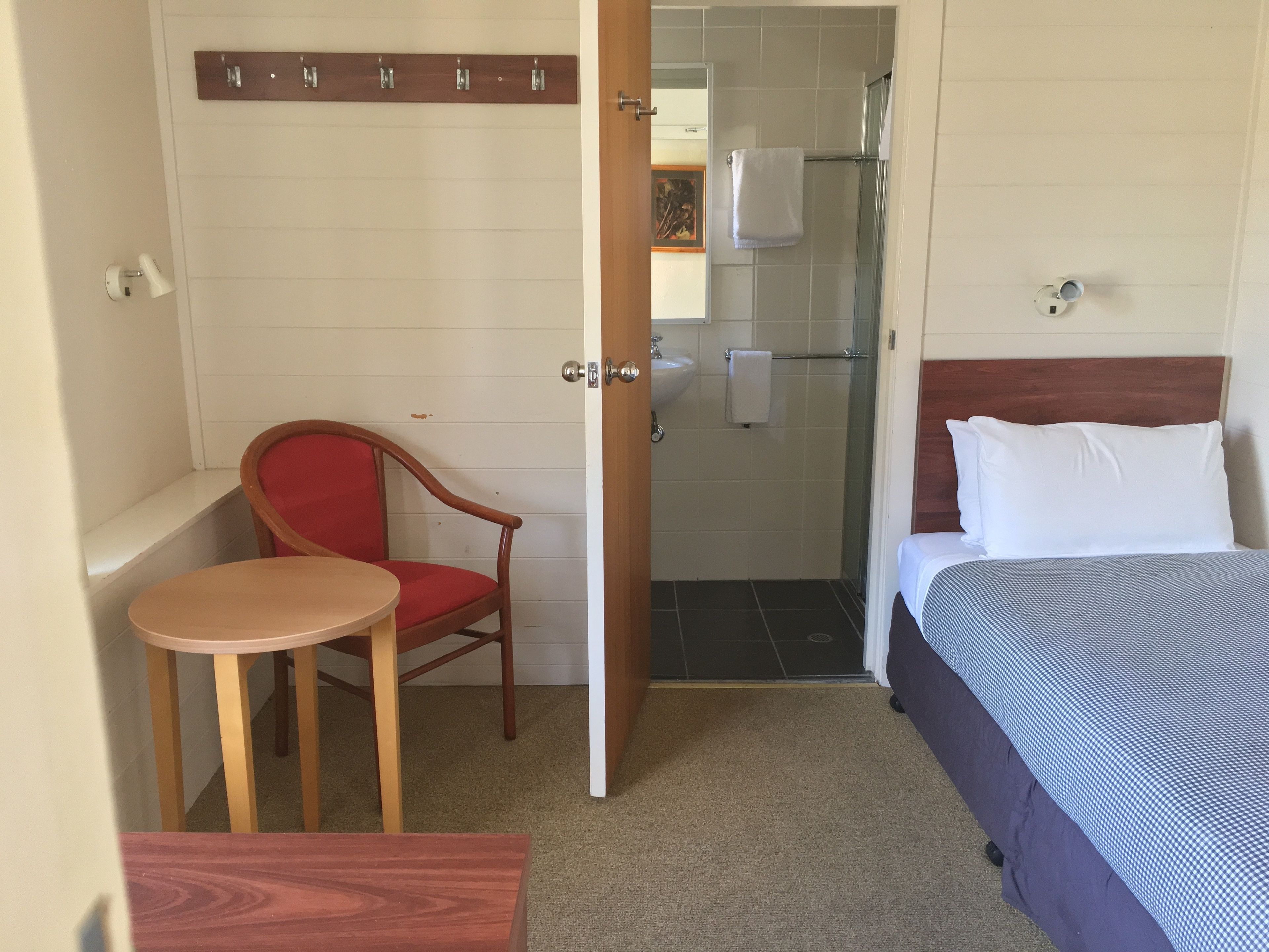 A small, compact room with a bed, a chair, a small table, a shower, and a wooden coat rack.
