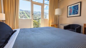 A hotel room with a large bed, a chair, a lamp, and a window with a view of greenery. - The Denman Hotel (Thredbo)