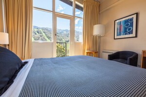 Quarto de hotel com uma cama grande, uma cadeira, um abajur e uma janela com vista para o verde.