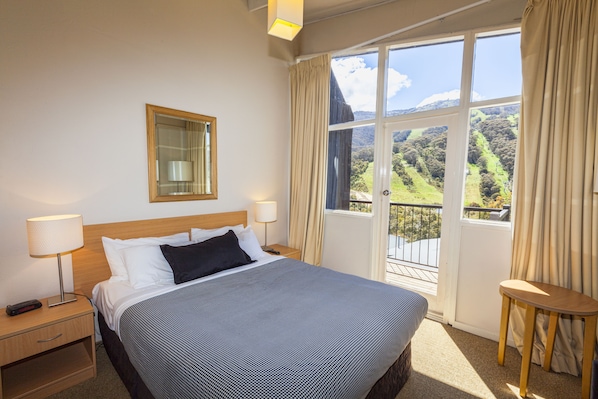 A hotel room with a large bed, two bedside tables, a nightstand, a mirror, and a view of a green landscape through a large window. - The Denman Hotel (Thredbo)