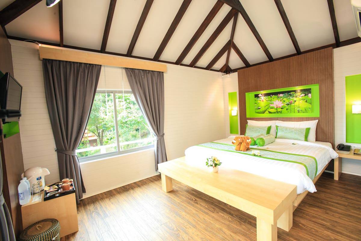 A bedroom with a wooden ceiling, a large bed with green and white bedding, a bedside table with a lamp, and a window with curtains.