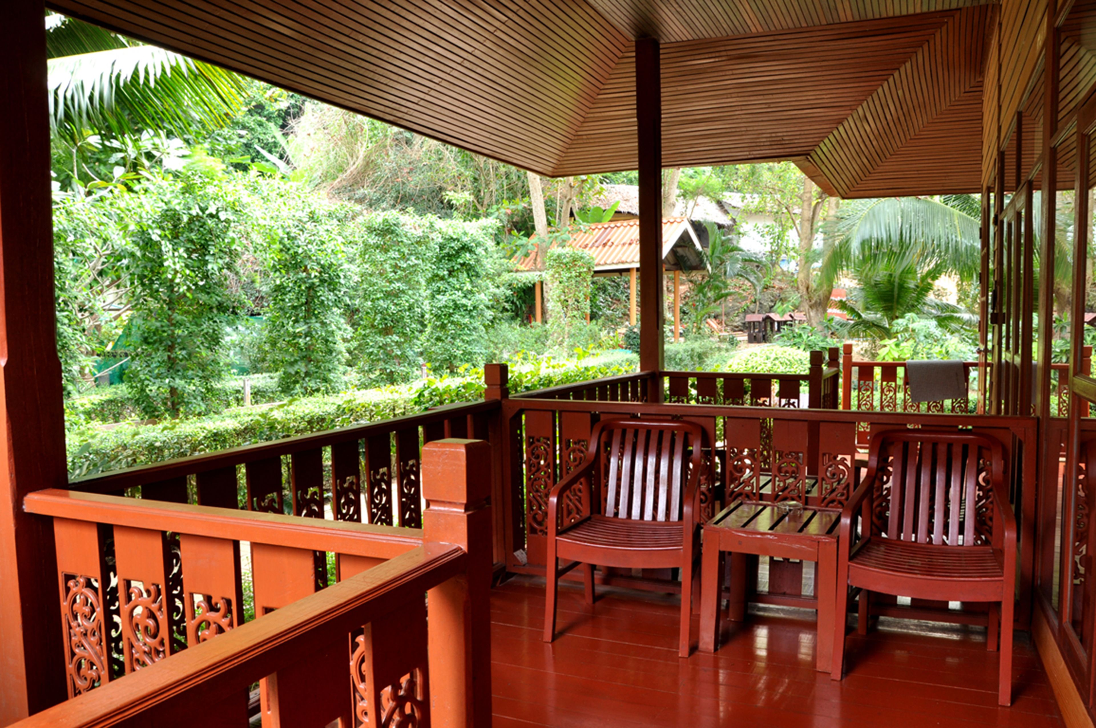 A wooden deck with two chairs and a table, surrounded by lush greenery and a traditional house in the background.