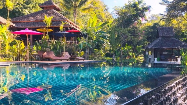 A clear blue infinity pool with lounge chairs and umbrellas, surrounded by lush greenery and traditional thatched-roof structures. - Fern Resort (Mae Hong Son)