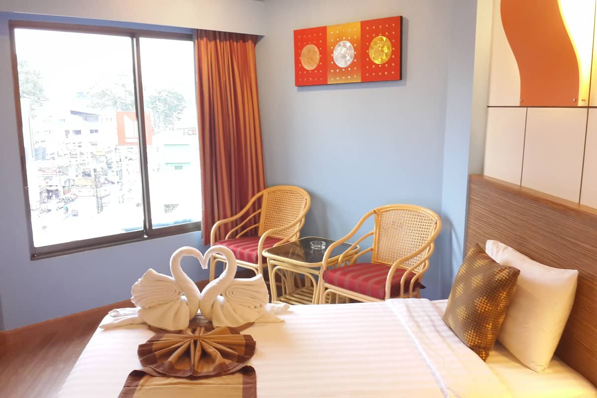 A hotel room with a bed, two wicker chairs, a table, a view of the street through a window, and a wall decoration.
