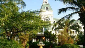 A formal garden with a fountain, palm trees, and a building with a green roof and arched windows. - Camelot Hotel Pattaya (Pattaya)