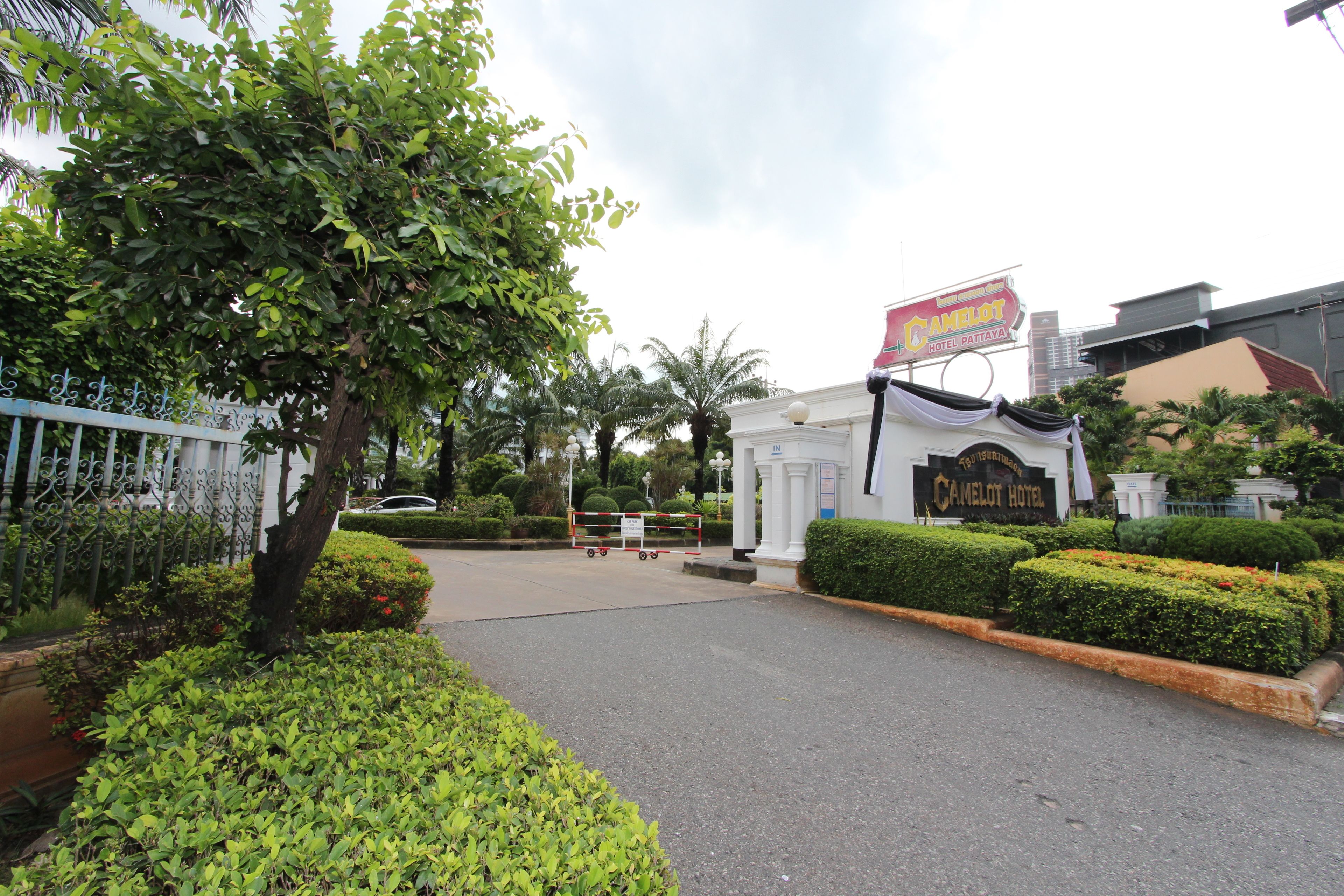 A gated entrance to Camelot Hotel with a sign, surrounded by well-maintained hedges and a tree.