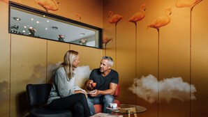 A room with a wooden wall, a mirror, and a wall mural of flamingos. There are two chairs, a small round table, and a magazine on the table. - HEINhotel Vienna Airport (Schwechat)
