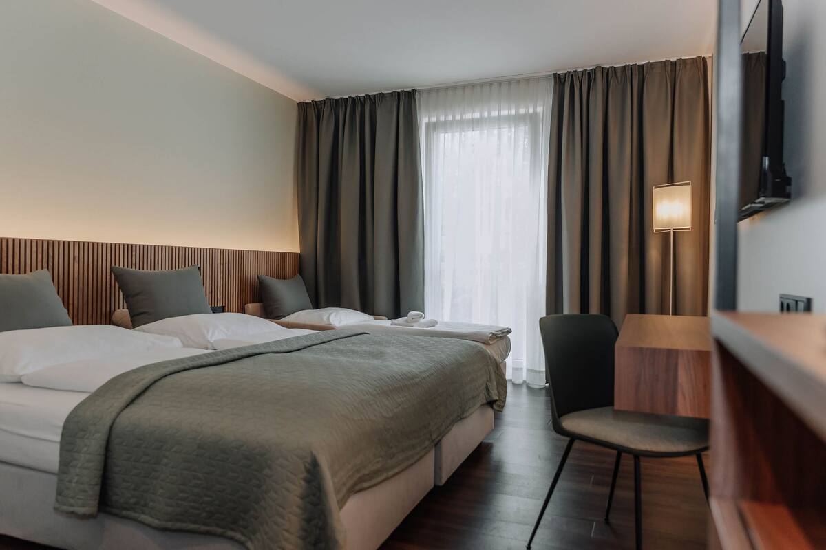 A hotel room with two beds, a desk, a chair, a TV, and a window with curtains.