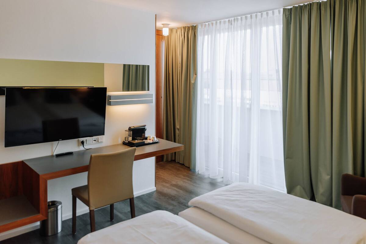 A hotel room with a flat-screen TV, a desk with a chair, a bed, and a window with curtains.