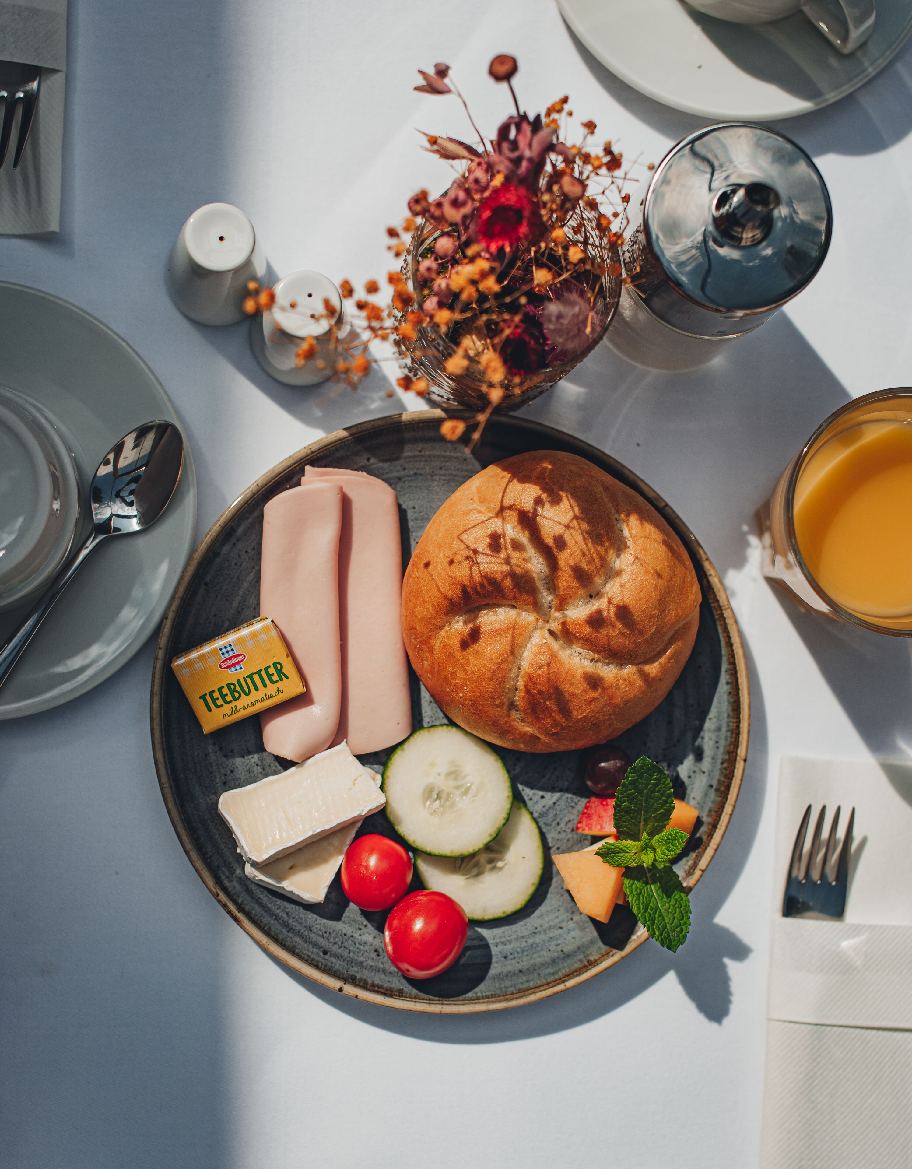 Un desayuno con un plato de jamón, queso, pepino, tomates cherry y un muffin, junto con un vaso de zumo de naranja y un jarrón de flores.