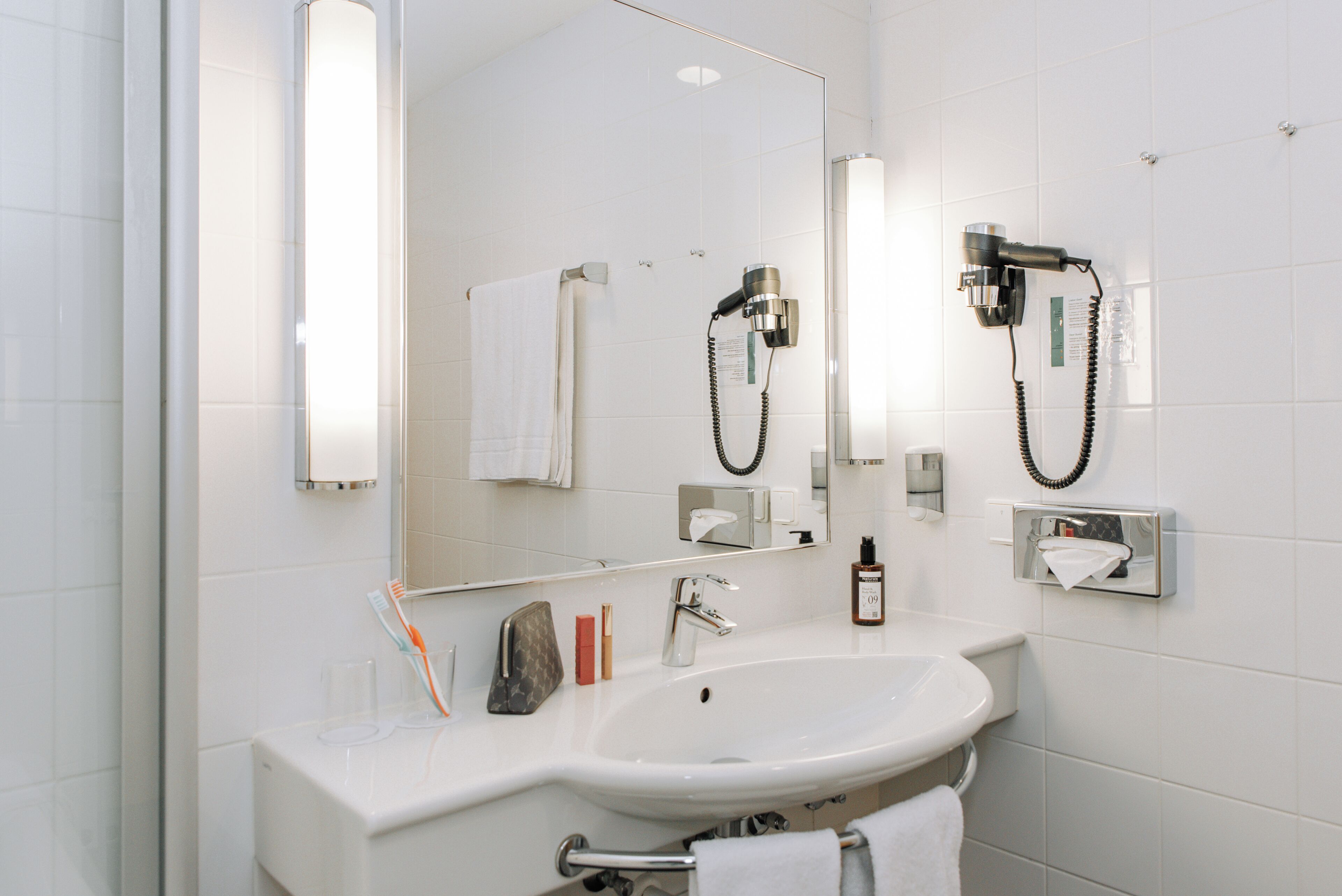 Ein modernes Badezimmer mit einem weißen Waschbecken, einem großen Spiegel, einem an der Wand montierten Föhn und einem Handtuchhalter.