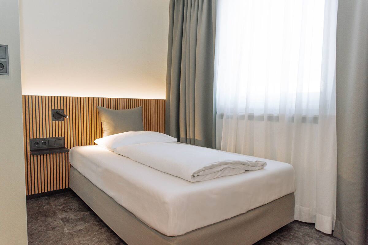 A neatly made bed with a pillow, a bedside table with a lamp, and a window with curtains.