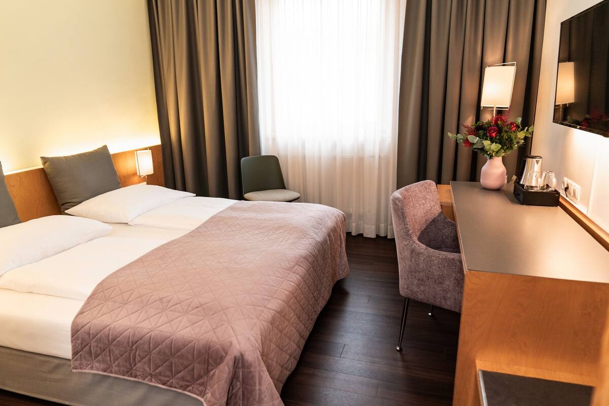 A hotel room with a large bed, a desk with a lamp, a chair, and a vase with flowers.