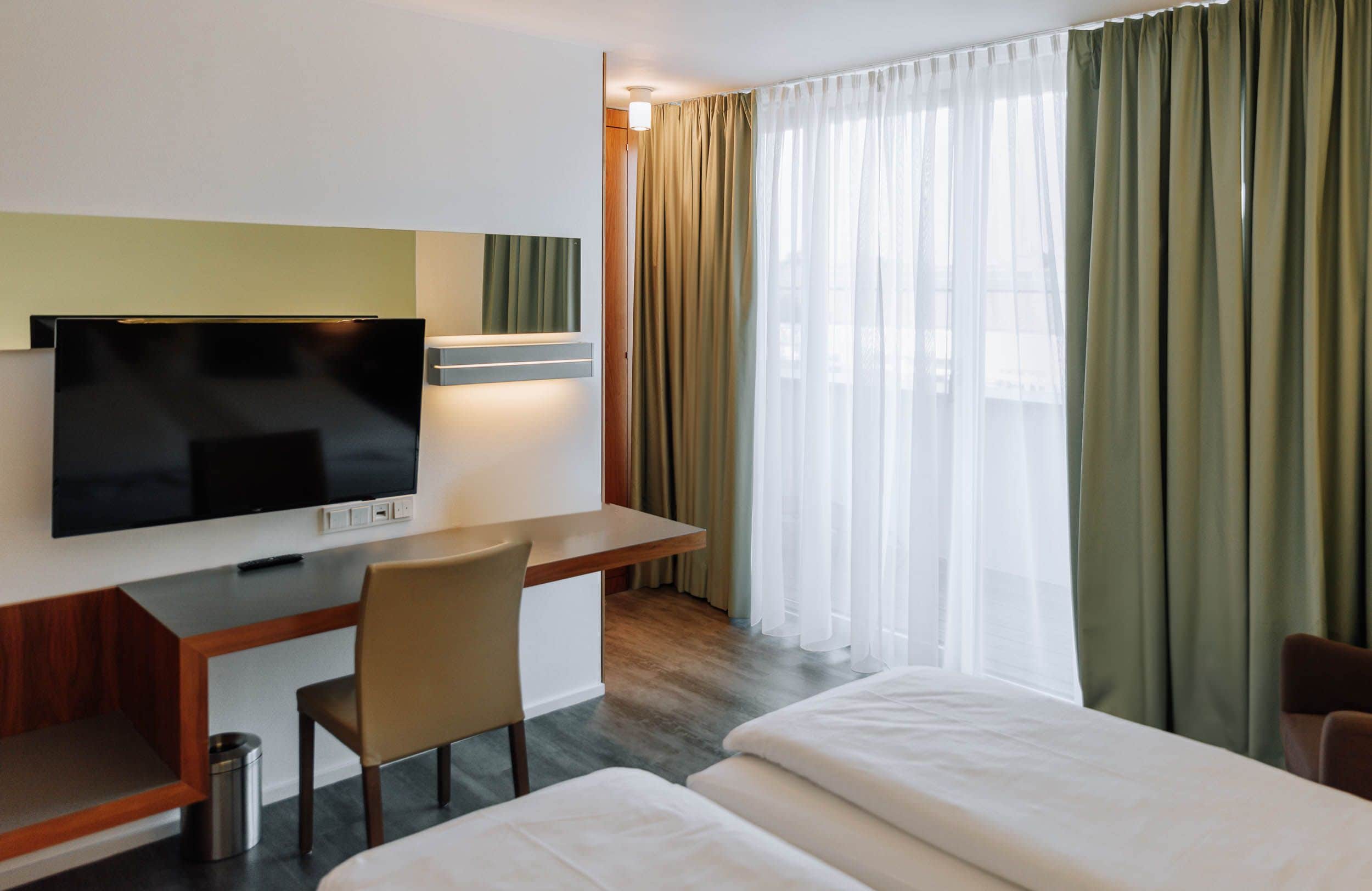 Una habitación de hotel con cama, escritorio con televisor, silla y ventana con cortinas.